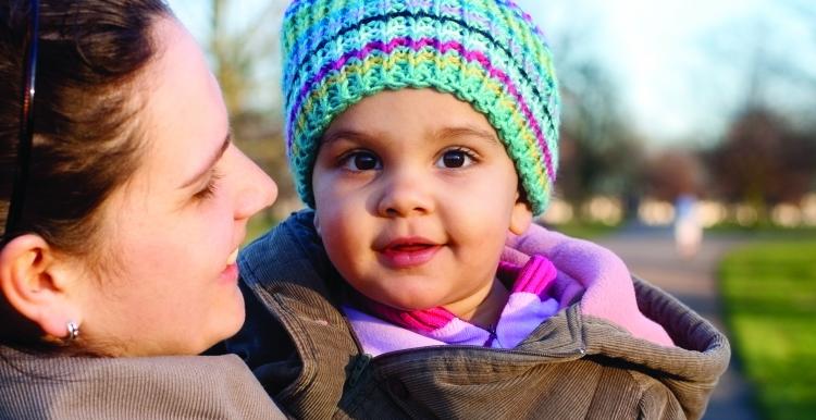 A baby in a knitted hat is held by her mum and looks at camera 