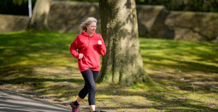 a woman with grey hair running through a park. she is wearing a red hoodie and black leggings. 