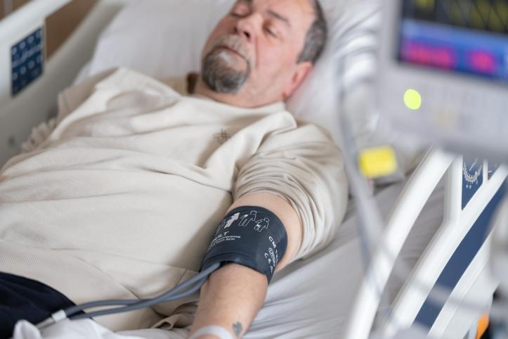 A man with a grey beard, wearing a beige t shirt lying in a hospital bed with his eyes closed. He has a blood pressure cuff on his arm.