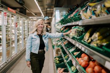 A woman with long blonde hair is standing in a supermarket with one hand in the fruit and veg aisle, and the frozen section to her left