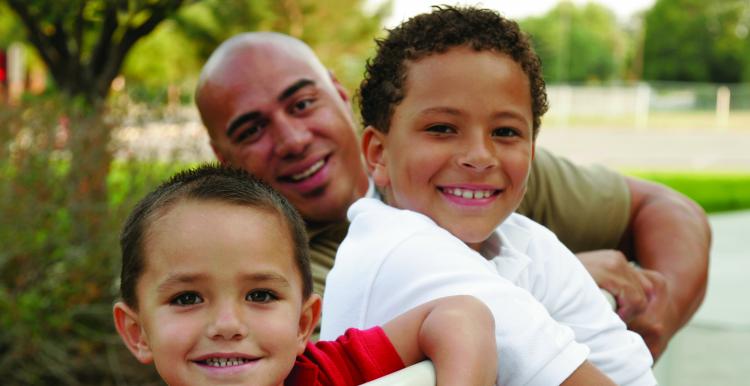 Two boys lean over a fence outside with their Dad