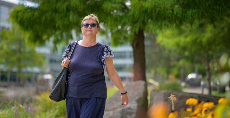 A woman wearing grey short-sleeved top and trousers is walking outside. Yellow flowers are on the right hand side and a green tree is in the background. The woman is wearing sunglasses.