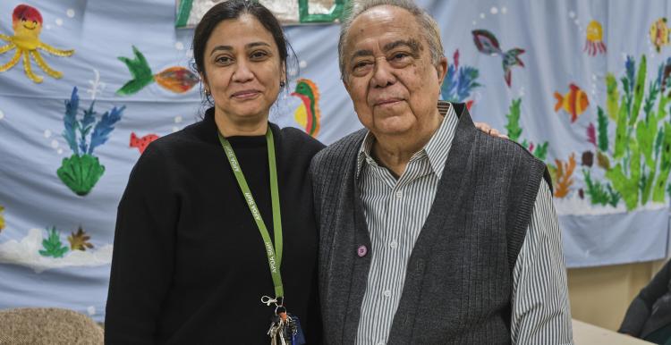 An Asian woman and older man stand side by side. She is wearing a black jumper with a lanyard around her neck, he is wearing a grey shirt and knitted waistcoat. The background behind them is bright blue with hand painted flowers.