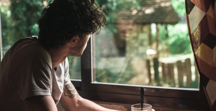 A young person looking out of a window, wearing a t shirt, and a glass and plate in front of him on a table