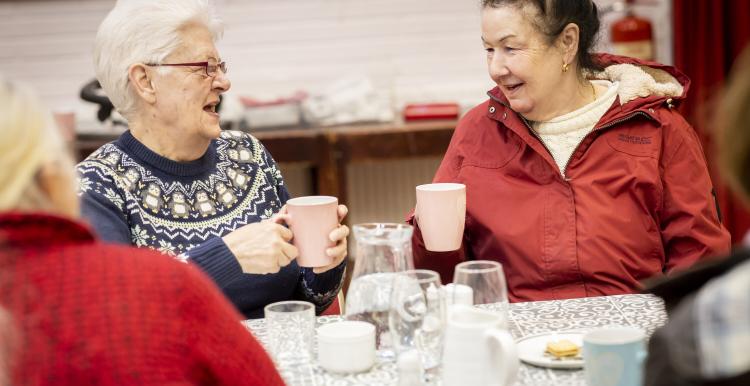 Two older women sit at a table. Both are holding a mug, they are talking. One has white hair and a blue jumper, the other had brown hair and a red coat. 