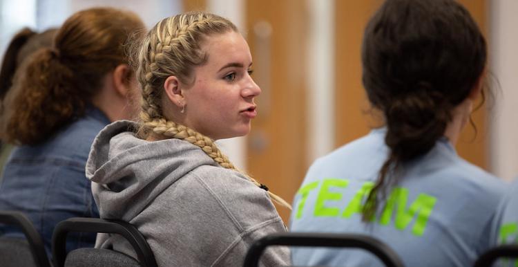 Three young girls with their backs to the camera, on the left a girl with blonde hair in plaits and a grey hoodie is talking to another girl on her right hand side, who is wearing a light blue t shirt.