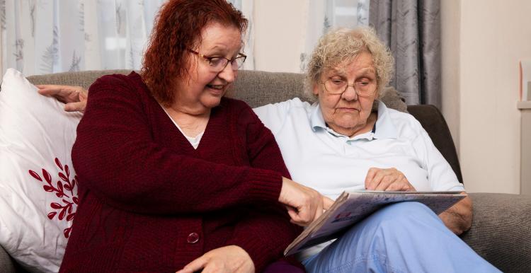 Two older women sitting on a sofa, one wearing a maroon cardigan and pointing at something in a newspaper