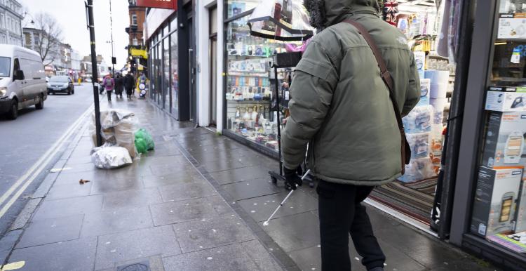 A person wearing a coat with their hood up and walking along a busy street lined with shops. It looks cold and its raining.