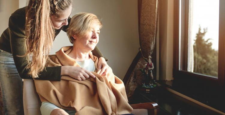 A woman is wrapped in a blanket and sits by a window with her carer 