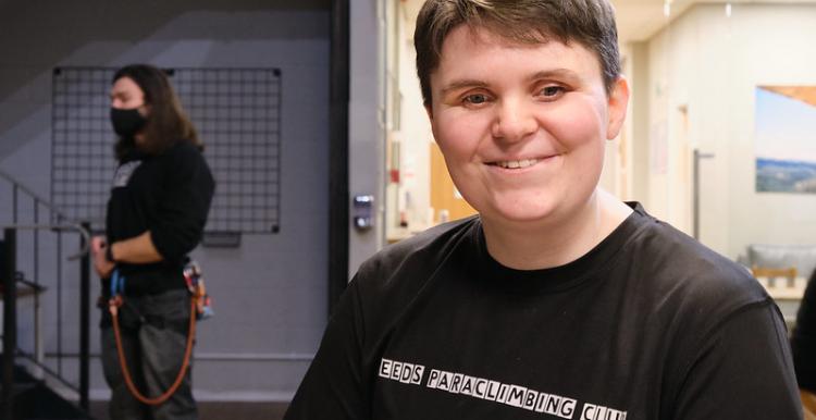 A woman who is blind wearing a black t shirt smiles at the camera. She is at a climbing club. 