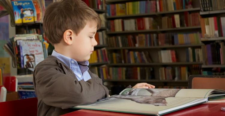 A boy surrounded by book shelves sitting down looking at a book 