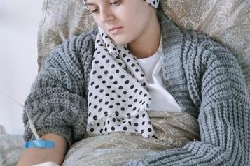 A woman wearing a head scarf and a grey cardigan, sitting in bed holding her phone with a medical tube in her arm