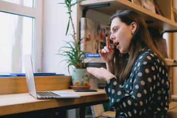 A woman with brown sitting by a window in front of a laptop, using sign language hand gestures. She is wearing a balck and white polka dot top, and a green plant sits on her desk