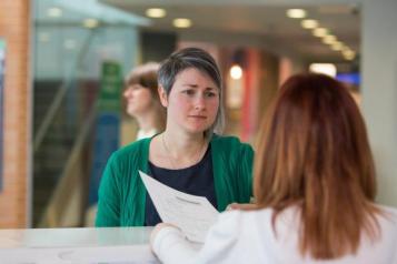 A woman with short hair and a green cardigan standing at a doctor's reception desk