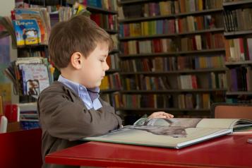 A boy surrounded by book shelves sitting down looking at a book 